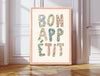 Floral Bon Appetit kitchen print with pastel flower letters and scalloped peach border, leaning against a white panelled wall