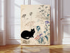 Japanese style print of a black cat watching a white barn owl among ink-drawn wildflowers, on a large canvas leaning against a white panelled wall