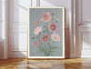 October birth flower art print of pink cosmos flowers on dusty blue in a gold frame, Cosmos October lettering, leaning against a panelled wall