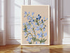 Larkspur botanical birth flower print resting on a wicker chest beside a bed and olive tree in a neutral bedroom